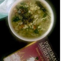 Green cabbage kootu/ stew with lentils and coconut paste, in a white bowl, garnished with coriander leaves. A red and black book by a Tamil author in the foreground, all on a black backgroundand a white and green napkin at the back.