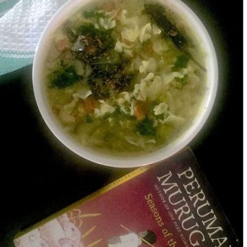 Green cabbage kootu/ stew with lentils and coconut paste, in a white bowl, garnished with coriander leaves. A red and black book by a Tamil author in the foreground, all on a black backgroundand a white and green napkin at the back.