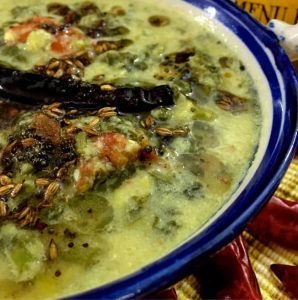 Green amaranthus stew with lentils and coconut paste in a green edged bowl, tempered with dry red chili, mustard and cumin seeds. Dry red chilies seen on the side, resting on a yellow straw mat