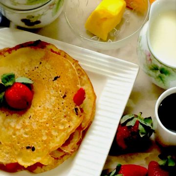 Really Easy Strawberry Buttermilk pancakes on a white square edged plate with a strawberry on top, syrup, milk and butter in little flowered cups alongside