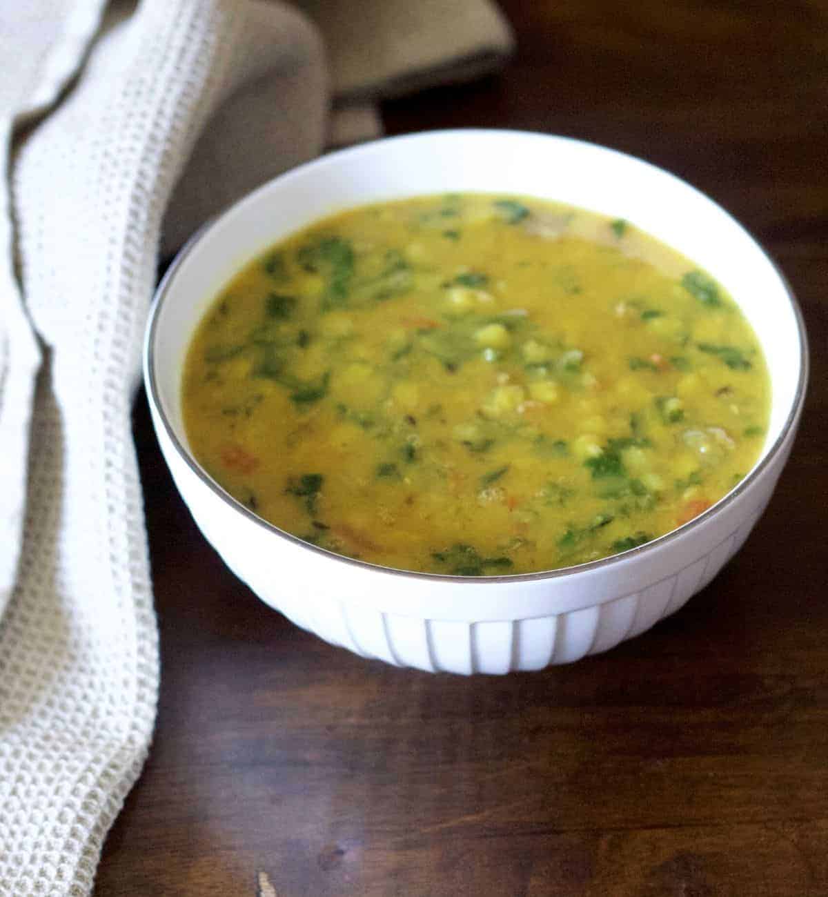 Yellow Arhar Dal palak or split yellow lentisl with green spinach seen in a white bowl. A pale grey napkin on the left, all on a dark brown base