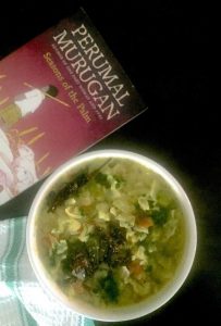 Easy green cabbage stew with vegetables, lentils and coconut, in a white bowl. Tempered with mustard, chili, cumin in coconut oil and garnished with green coriander. A pink and red book by Perumal Murugan at the back, all on a black background