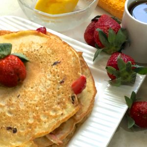 A stack of strawberry pancakes on a square white plate with plum strawberries scatter at one side and butter, milk and syrup in little glass or white bowls alongside