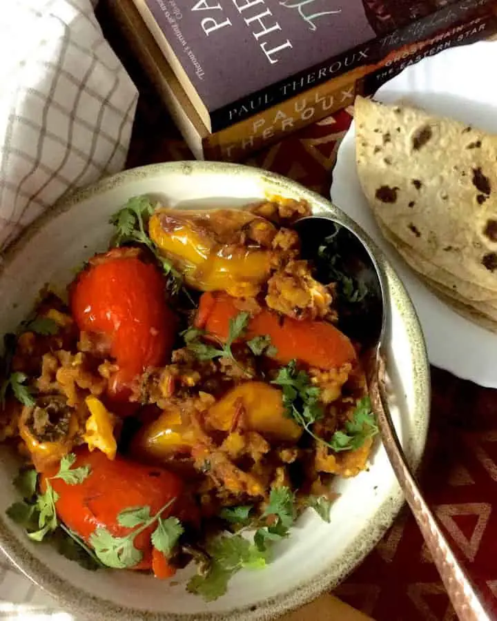 A round plate of stuffed Shimla mirch or coloured bell peppers stuffed with potato mash with a plate of chapatis, books and a checked napkin on the side