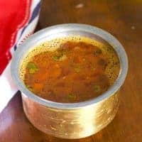 Brass vessel filled with reddish brown Tomato rasam. Pieces of tomato floating on top. A red and white napkin to the left, all of it on a brown wooden background