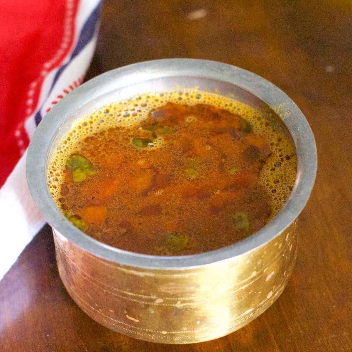 Brass vessel filled with reddish brown Tomato rasam. Pieces of tomato floating on top. A red and white napkin to the left, all of it on a brown wooden background