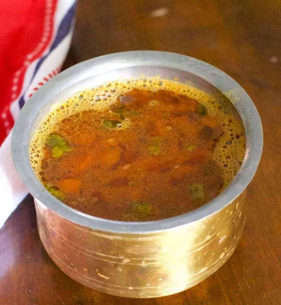 Brass bowl with straight sides and a rim, filled with reddish brown liquid with slices of tomato and green coriander leaves floating on top. A red and white napkin to the left, on a wooden background