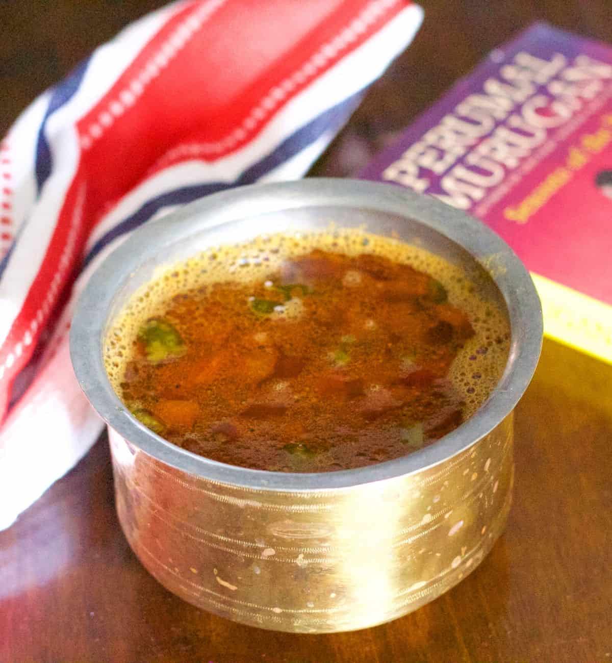 Reddish brown rasam in a brass vessel on a brown wooden background. Tomato slices seen floating on top. A red black and white napkin to the left and a book by Tamil author Perumal Murugan to the right, with a pink red cover