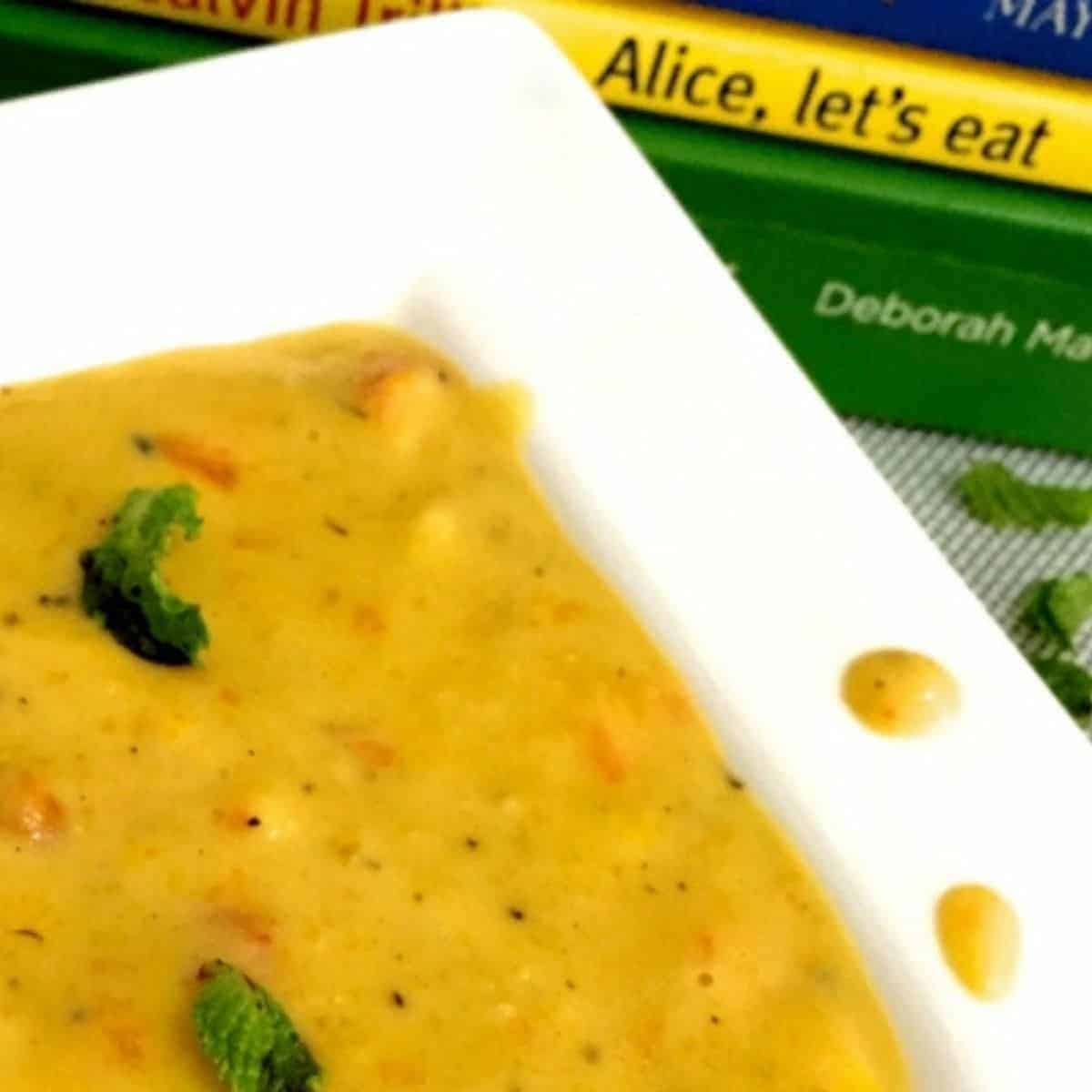 A broad white edged plate iwth yellow orangey vegetable corn chowder, garnished with mint leaves and flecked with black pepper. Green and yellow food books seen in the background