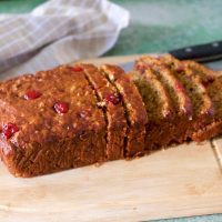 Red brown Banana Bread studded with bright red cranberries. sliced and placed on a light wooden cutting board. A pale grey cheked napkin behind, and a black knife handle seen to the right. All on a green background