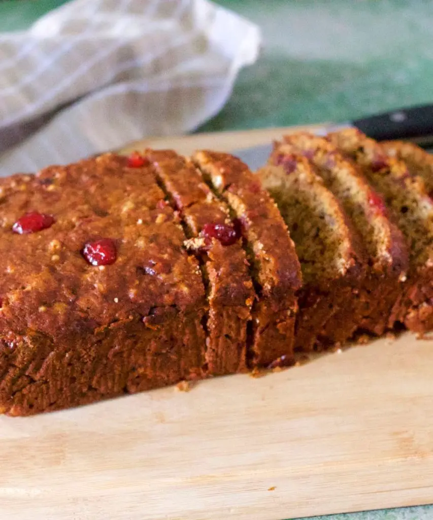 Loaf of reddish brown Banana bread with cranberries, sliced on a light wooden cutting board. A grey and white checked napkin and a black handled steel knife at the back, on a green background
