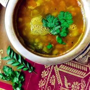 A brass bowl with soup like Pineapple Rasam garnished with coriander leaves, tempered with mustard and cumin in ghee, resting on a red slik fabric embroidered with gold thread. Curry leaves and coriander leaves at the bottom left