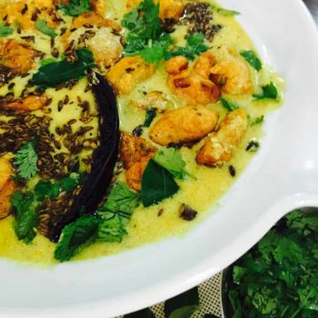 White rimmed bowl with yellow yogurt based kadhi with chickpea fritters and tempering of cilantro, cumin and red chilies