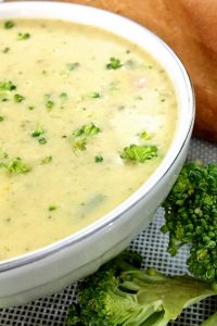 Creamy Yellow broccoli cheddar soup flecked with green bits of broccoli and herbs in a white bowl with a piece of green broccoli at the side and a piece of baguette in the backgroun