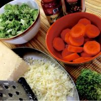 Ingredients for broccoli cheddar soup. Sliced carrots in an orange bowl, broccoli florets in a white bowl edged with gold, a plate with a grater and grated cheese