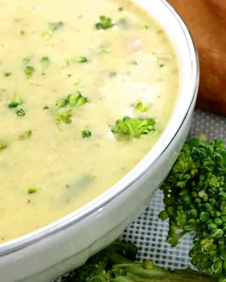 Yellow Broccoli Cheddar Soup flecked with green bits of herbs and broccoli,in a white bowl with a baguette in the background and pieces of green broccoli on the side