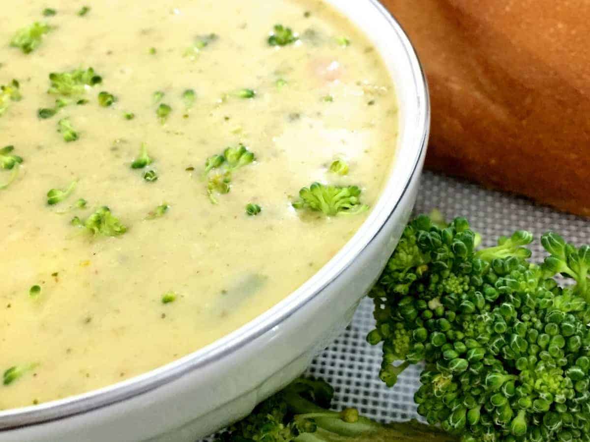 Yellow Broccoli Cheddar Soup flecked with green bits of herbs and broccoli,in a white bowl with a baguette in the background and pieces of green broccoli on the side