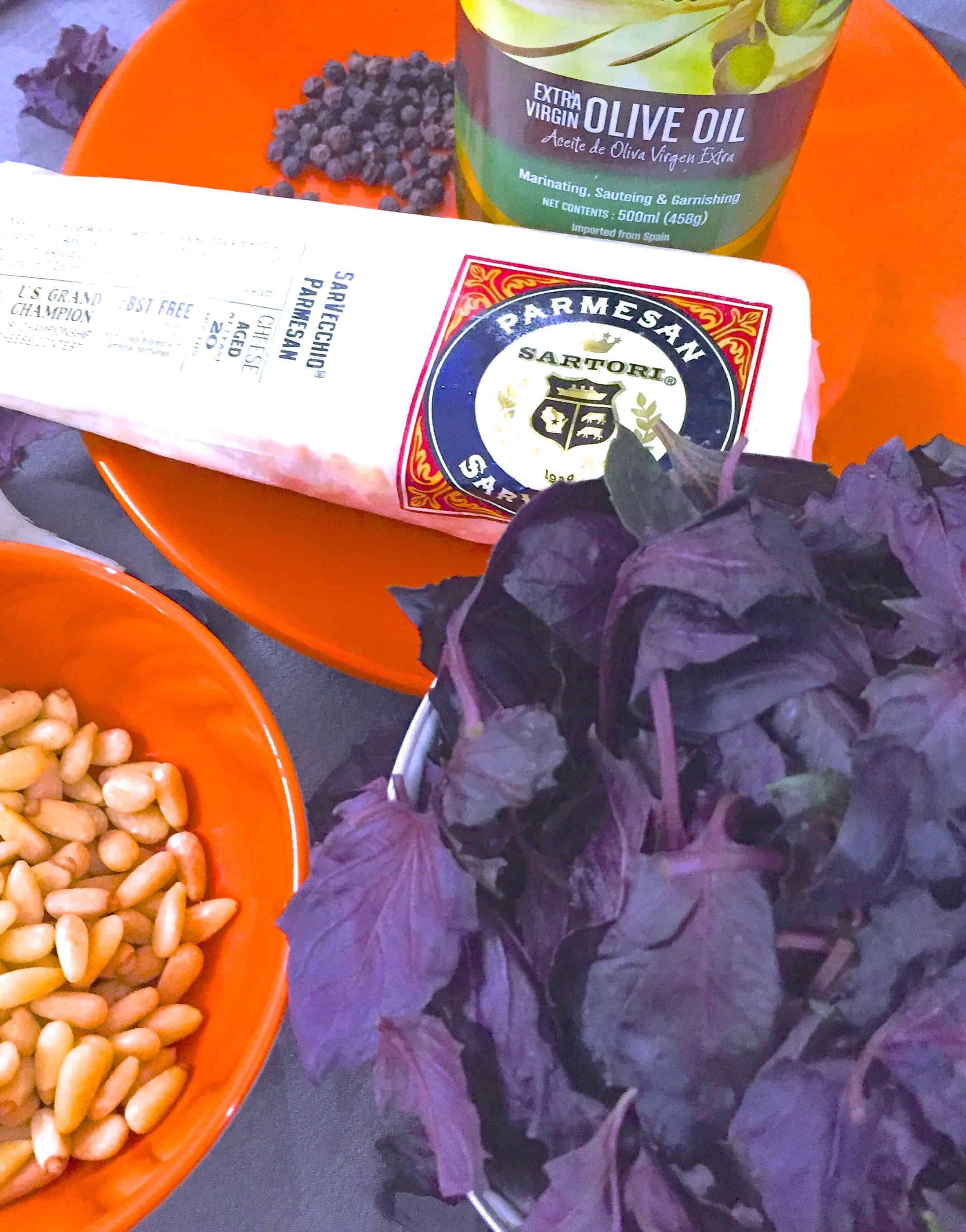 Ingredients for making purple basil pesto. Dark purple basil leaves with a slab of parmesan, black peppercorns and virgin olive oil on an orange plate and pine nuts on an orange bowl