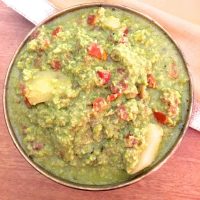 Green hued Green Pea curry with red tomato and chunks of potato showing through, in a round brass bowl. On a brick coloured background with a beige fabric in the background