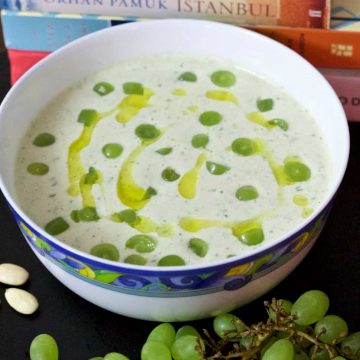 Chilled White Grape and Almond Gazpacho in white bowl edged with blue and yellow pattern, with green grapes and olive oil garnish. Green grapes and almonds on the foreground and books at the back including Istanbul by Orhan Pamuk