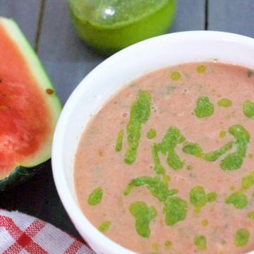 Pink Watermelon Gazpacho garnished with homemade Basil Oil, in a white bowl with a slice of watermelon on the left, a red checked napkin below and bottle of green basil oil in the background