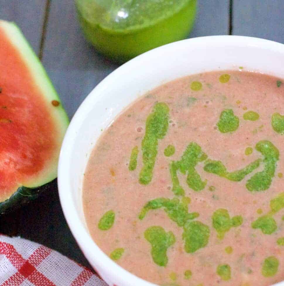 Pink Watermelon Gazpacho garnished with homemade Basil Oil, in a white bowl with a slice of watermelon on the left, a red checked napkin below and bottle of green basil oil in the background