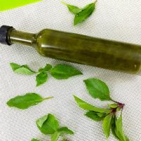 Homemade Basil Oil in a long narrow bottle, with basil leaves strewn around, on a white background edged with green