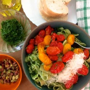 Fettucine in arugula pesto with burst cherry tomatoes ladled on top. A bowl of Pistachio nuts and a glass bowl with arugula pesto and a bottle of olive oil on the left and a plate of bread and a green white checked napkin on the side