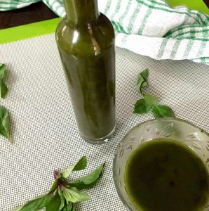 A tall narrow bottle filled with dark green basil oil, on white green edged plastic sheet, with basil leaves strewned around and a glass bowl of basil oil in the foreground. A white an green striped napkin in the background