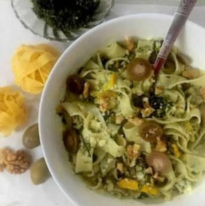 Fettucine fennel frond pesto in a white bowl, garnished with watlnuts and olives. Some fettucine pasta balls, walnuts and olives on the left side of the bowl and a glass bowl with pesto in the background