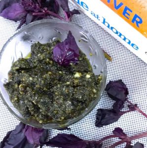 Green coloured Pesto of Purple basil leaves in a glass bowl. Purple basil leaves scattered around and books by Jamie Oliver, one with a mustard colour and another white, in the background