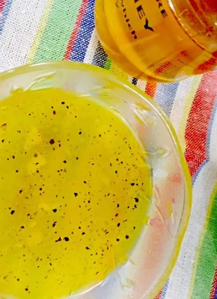 A glass bowl with yellow garlickly honey lemon salad dressing, speckled with black pepper Powder. A small bottle of golden honey seen in the background. All on a rainbow striped napkin