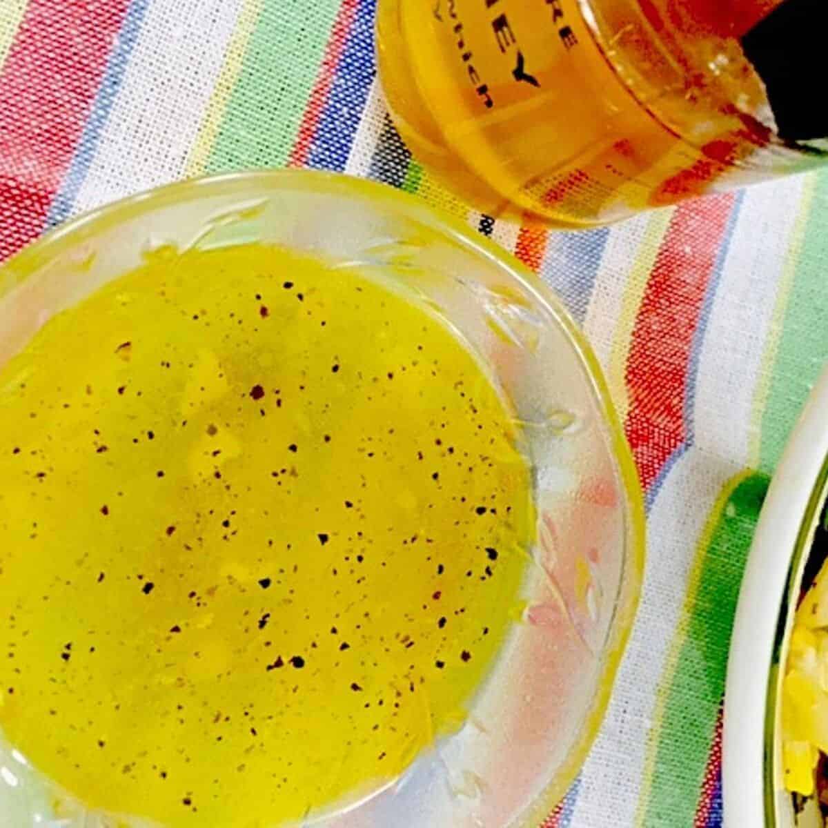 A glass bowl with yellow garlickly honey lemon salad dressing, speckled with black pepper Powder. A small bottle of golden honey seen in the background. All on a rainbow striped napkin