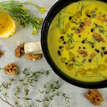 Yellow turmeric walnut fennel soup in a large cup with a handle on a white mat with yellow flowers and walnuts strewed alongside