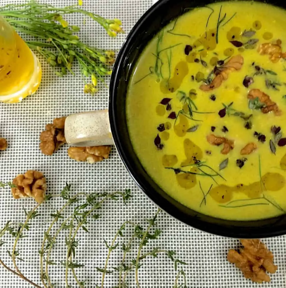 Yellow turmeric walnut fennel soup in a large cup with a handle on a white mat with yellow flowers and walnuts strewed alongside