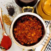 A brass bowl with fiery red spicy raw mango thokku, a South Indian relish of mango slices cooked in turmeric, chili, oil, spices. A brass spoon with red chili powder to the left, fenugreek and mustard seeds top left and another brass spoon on a dark blue and white lined napkin to the right