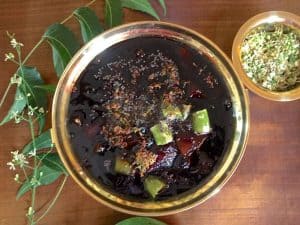 Brass bowl with almost black coloured mango pachadi, granished with green chili, mustard and fried neem blossoms. A twig of neem leaves and flowers to the left