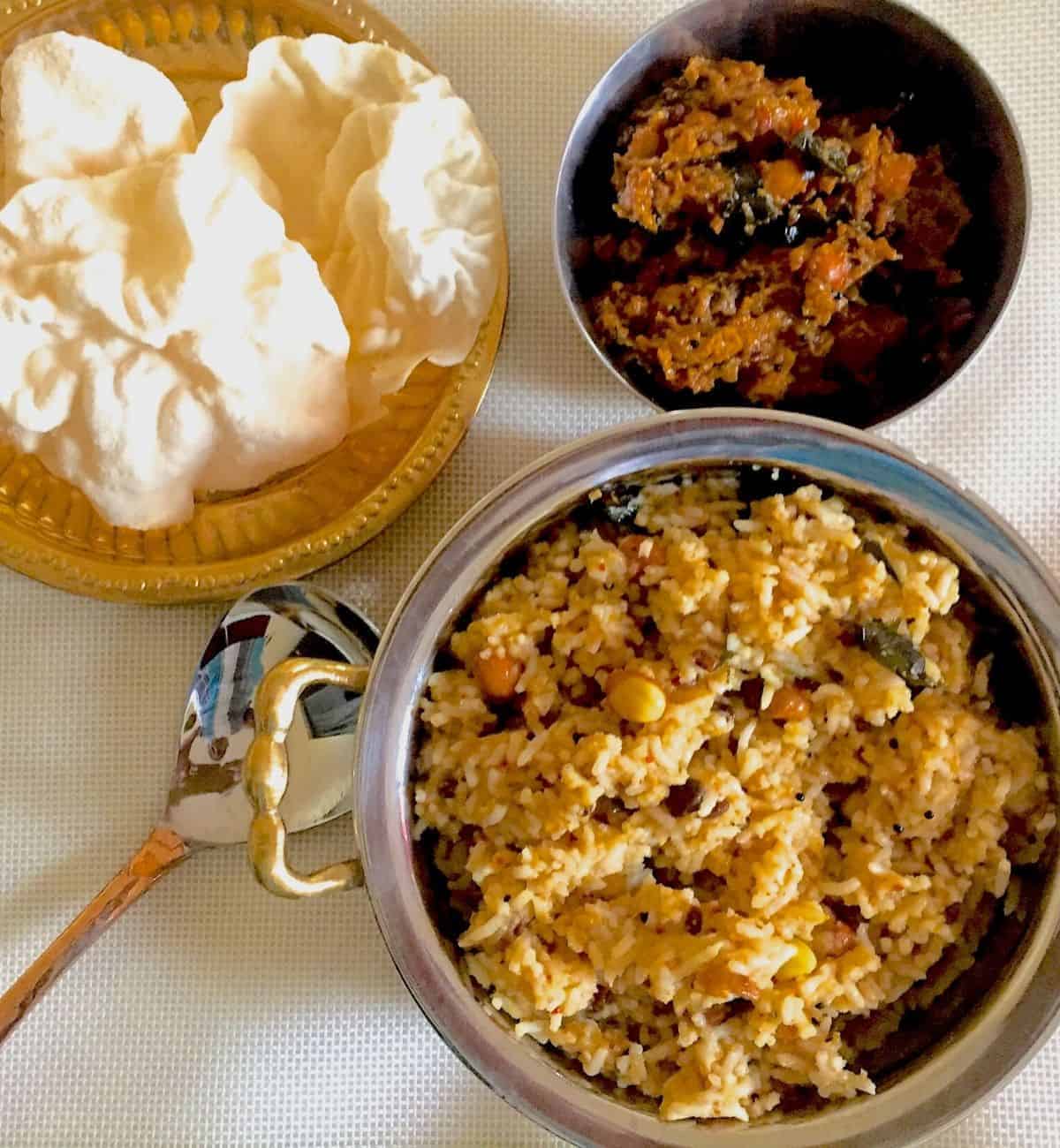 Light brown Puliyodharai Tamarind Rice with peanuts in a brass bowl along with a plate of fried pappad and a bowl of brown tamarind paste. A spoon lies between the bowls which are all on an off white background