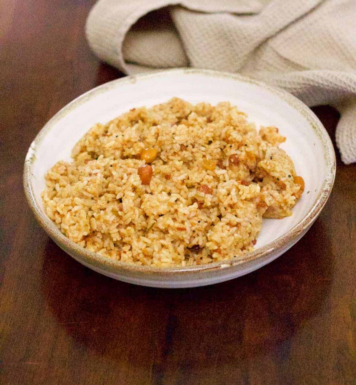 Brown hued Puliyodharai or tamarind rice in a white bowl edged with beige, on brown wooden background. A beige napkin lies behind the bowl