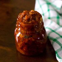 Glass jar heaped with reddish brown raw mango thokku, a pickle like relish. On a brown background with a white and green checked napkin to the right