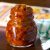 Glass jar filled with reddish brown mango relish in oil. On a brown background. A yellow, white and pink covered book in the background and a white and green checked napkin by the side