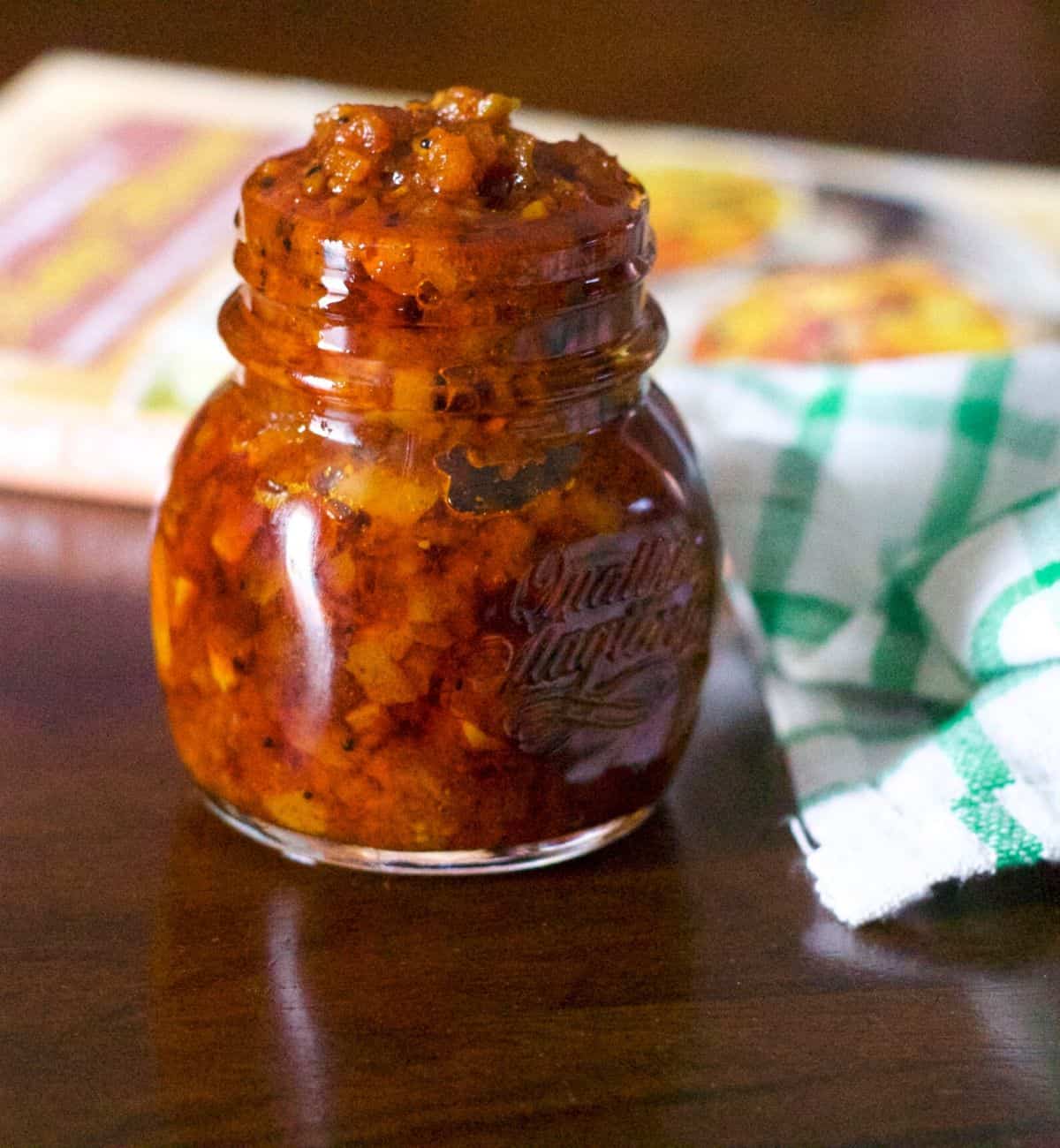 A small glass jar filled with a red brown relish or Raw Mango Thokku. On a brown background with a yellow and white covered book in the background and a white and green napkin by the side