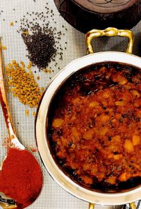 Large brass bowl with spicy red mango slices cooked in spices and oil. The Mango Thokku Relish has mustard seeds, fenugreek seeds on the left with a brass spoon filled with red chili powder