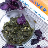 Purple basil in a glass bowl surrounded by scattered purple basil leaves, on a white background. Books by Jamie Oliver in the background