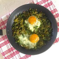 A pan of Green Shakshuka with Kale and Pesto with two open faced eggs, all on a red striped napkin on a white background