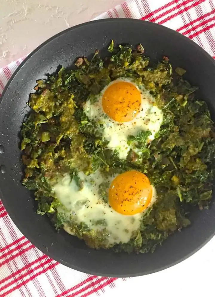 A pan of Green Shakshuka with Kale and Pesto with two open faced eggs, all on a red striped napkin on a white background