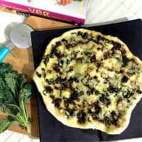 Thin crust kale and onion pizza on a black slab. A bunch of leafy kale on the left, a pizza cutter and a cook book with Pink and white binding in the background