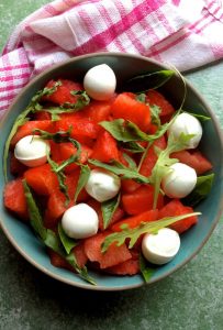 A round bowl with bright red watermelon with little spheres of bocconcini and fresh arugula leaves