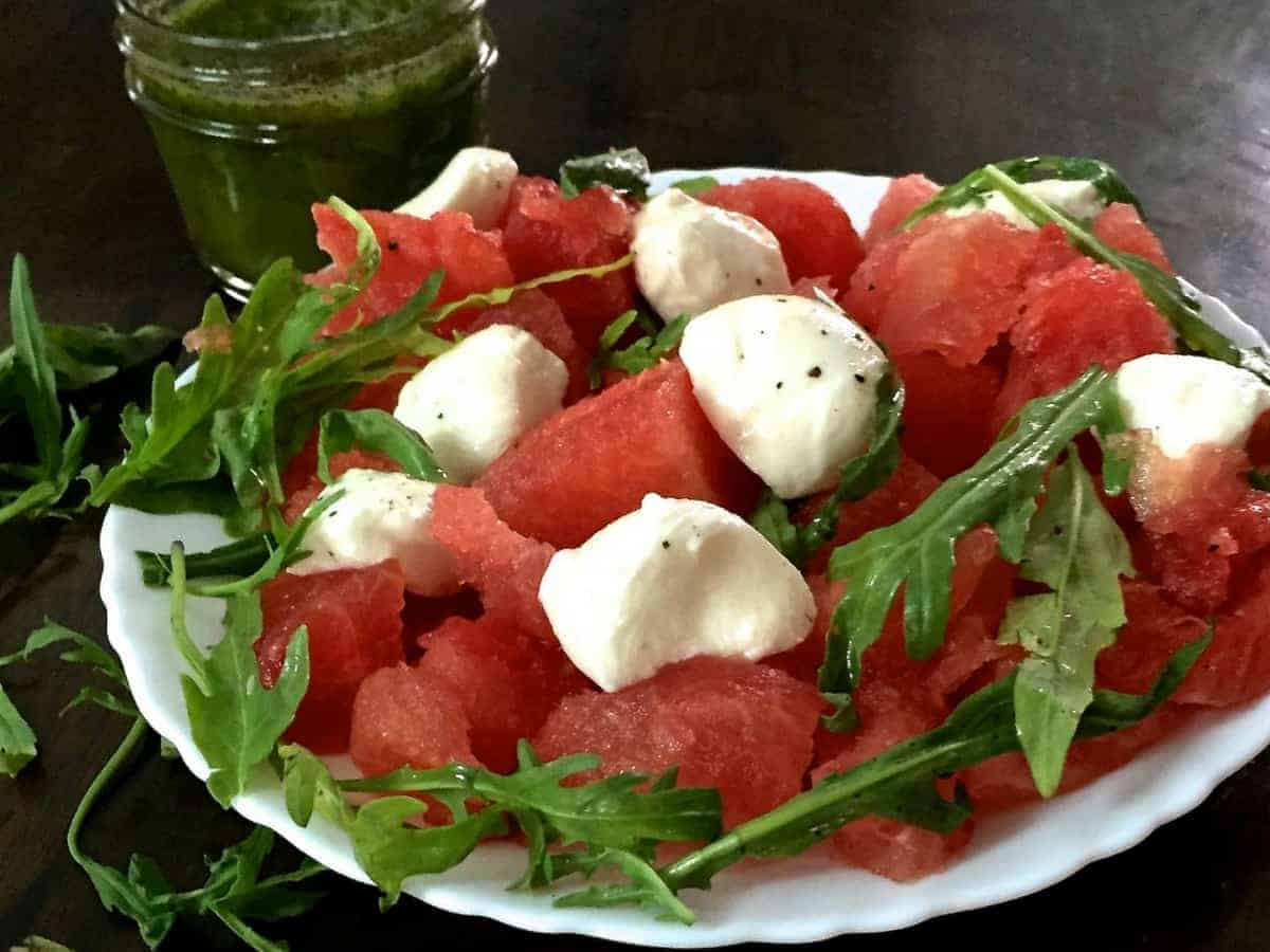 A white plate with a scalloped edge, filled with cubes of Watermelon and Halved Bocconcini cheese, with fresh arugual and pepper flakes for an easy cooling Watermelon Bocconcini Arugula Salad with Homemade Basil Oil