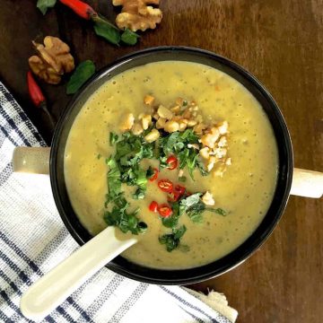 Overhead view of a cup of yellow curried chilli zucchini soup, garnished with slices of red chillies, coriander,parsley and mint leaves and walnuts. A white napkin striped with dark blue to the left bottom, and walnuts and red chillies top left